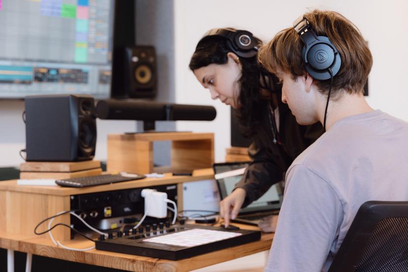 Two people work on audio production, using headphones and a sound mixer, in a modern studio with speakers and a screen.