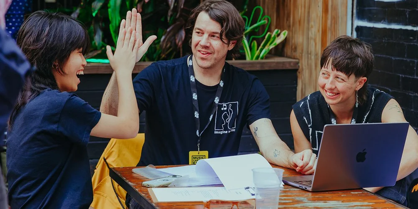 Three people sitting at a table outdoors, one giving a high-five, all smiling. A laptop and papers are visible on the table.